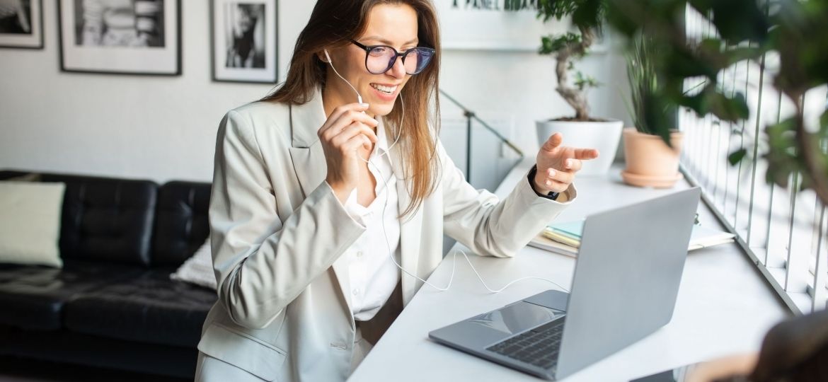 Positive female manager in earphones working on computer, consulting clients online, sitting in cozy coworking space